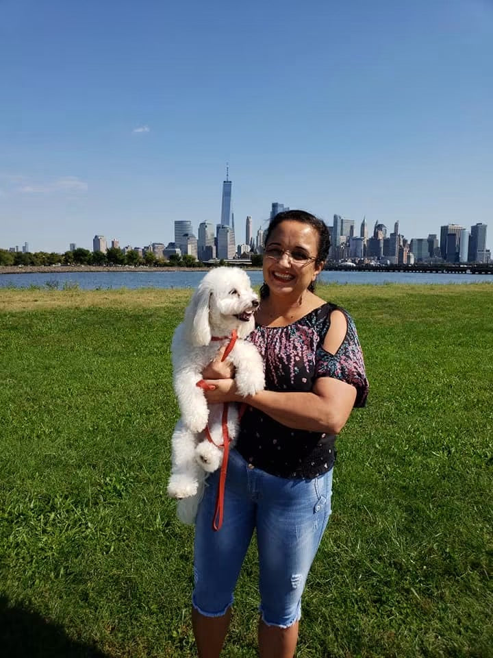 Geralda smiling while holding Luna in a park with the New York City skyline behind them.